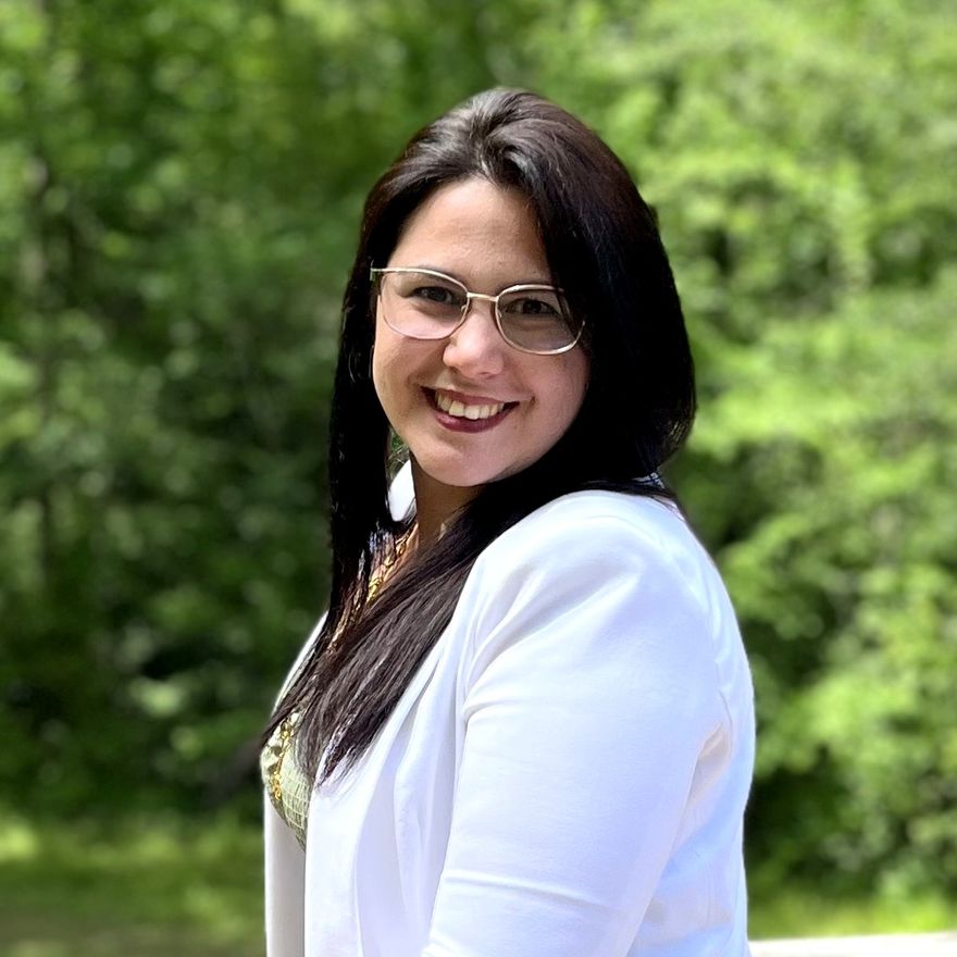 Retrato de Yainett Carbot Solis sonriendo, con chaqueta blanca y lentes, al aire libre.
